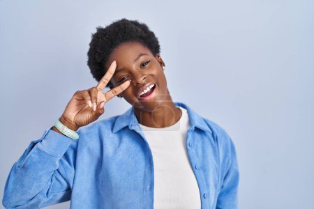 Téléchargez les photos : African american woman standing over blue background doing peace symbol with fingers over face, smiling cheerful showing victory - en image libre de droit