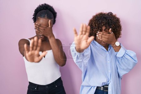 Téléchargez les photos : Deux femmes africaines debout sur fond rose couvrant les yeux des mains et faisant un geste d'arrêt avec expression triste et peur. concept embarrassé et négatif. - en image libre de droit
