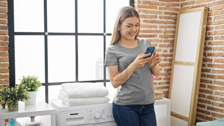 Foto de Mujer rubia joven usando smartphone esperando la lavadora en la sala de lavandería - Imagen libre de derechos