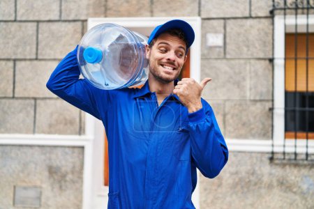 Foto de Joven hispano sosteniendo una botella de galón de agua para la entrega apuntando el pulgar hacia el costado sonriendo feliz con la boca abierta - Imagen libre de derechos