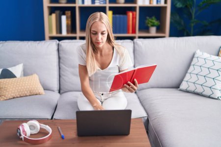 Photo for Young blonde woman student sitting on sofa reading book at home - Royalty Free Image