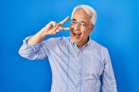 Foto de Hombre mayor hispano usando gafas haciendo símbolo de paz con los dedos sobre la cara, sonriendo alegre mostrando la victoria - Imagen libre de derechos