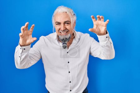 Téléchargez les photos : Homme d'âge moyen avec les cheveux gris debout sur fond bleu souriant drôle faisant griffe geste comme chat, expression agressive et sexy - en image libre de droit