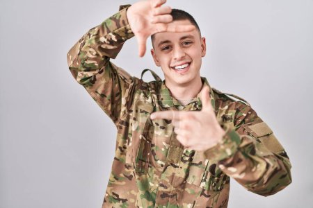 Téléchargez les photos : Jeune homme portant uniforme de camouflage de l'armée souriant faisant cadre avec les mains et les doigts avec le visage heureux. concept de créativité et photographie. - en image libre de droit