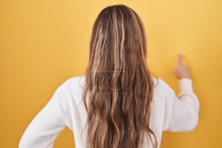 Téléchargez les photos : Jeune femme caucasienne portant un pull blanc sur fond jaune posant vers l'arrière pointant vers l'avant avec la main du doigt - en image libre de droit