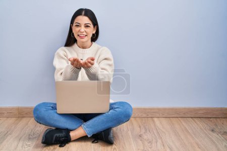 Téléchargez les photos : Jeune femme utilisant un ordinateur portable assis sur le sol à la maison souriant avec les mains paumes ensemble recevoir ou faire un geste. maintien et protection - en image libre de droit