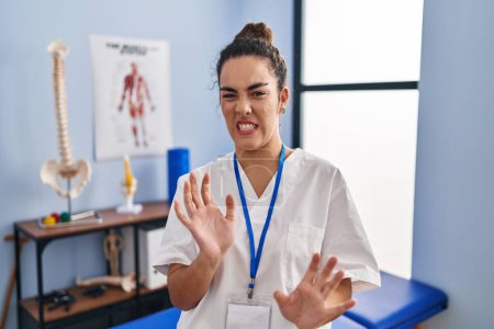Photo for Young hispanic woman working at rehabilitation clinic disgusted expression, displeased and fearful doing disgust face because aversion reaction. with hands raised - Royalty Free Image