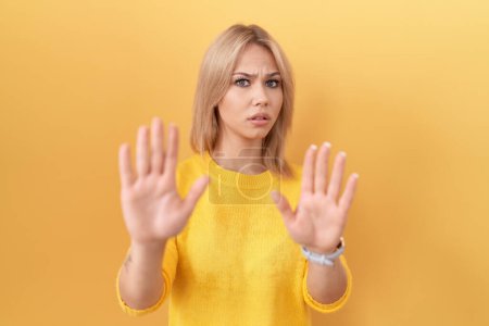 Foto de Mujer caucásica joven con suéter amarillo alejando las palmas de las manos mostrando rechazo y negación con miedo y expresión repugnante. parada y prohibido. - Imagen libre de derechos