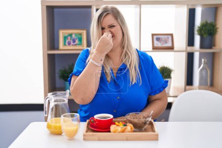 Foto de Mujer caucásica de talla grande desayunando en casa oliendo algo apestoso y asqueroso, olor intolerable, conteniendo la respiración con los dedos en la nariz. mal olor - Imagen libre de derechos