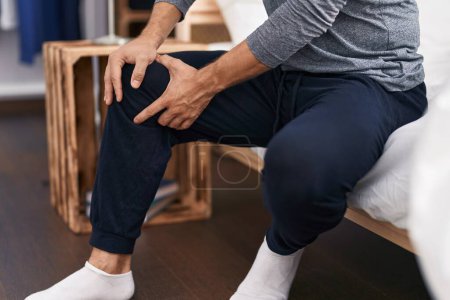 Téléchargez les photos : Young hispanic man suffering for knee injury sitting on bed at bedroom - en image libre de droit
