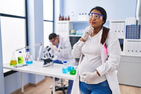 Photo for Young hispanic woman expecting a baby working at scientist laboratory serious face thinking about question with hand on chin, thoughtful about confusing idea - Royalty Free Image