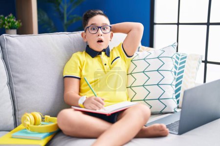 Photo for Young hispanic kid doing homework sitting on the sofa crazy and scared with hands on head, afraid and surprised of shock with open mouth - Royalty Free Image