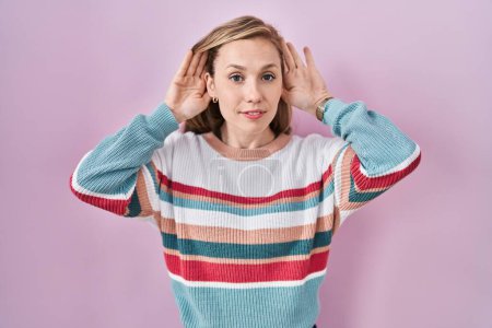 Téléchargez les photos : Young blonde woman standing over pink background trying to hear both hands on ear gesture, curious for gossip. hearing problem, deaf - en image libre de droit
