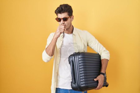 Foto de Hombre hispano joven sosteniendo maleta que va de vacaciones de verano sintiéndose mal y tosiendo como síntoma de resfriado o bronquitis. concepto de atención sanitaria. - Imagen libre de derechos