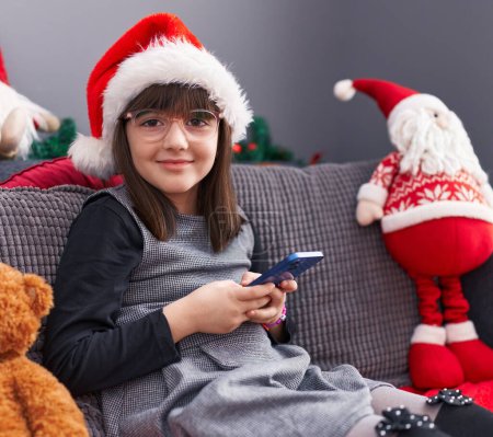 Téléchargez les photos : Adorable fille hispanique portant un chapeau de Noël en utilisant un smartphone à la maison - en image libre de droit