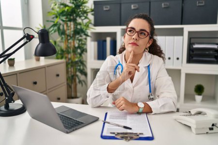 Photo for Young hispanic woman wearing doctor uniform and stethoscope thinking concentrated about doubt with finger on chin and looking up wondering - Royalty Free Image
