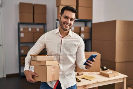 Photo for Young hispanic man e-commerce business worker using smartphone holding packages at office - Royalty Free Image