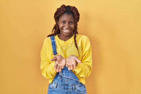 Foto de Mujer africana de pie sobre fondo amarillo sonriendo con las palmas de las manos juntas recibiendo o dando gesto. retención y protección - Imagen libre de derechos
