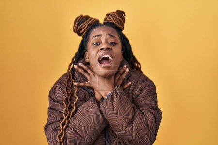 Foto de Mujer africana con el pelo trenzado de pie sobre el fondo amarillo gritando sofocada porque estrangulamiento doloroso. problema de salud. concepto de asfixia y suicidio. - Imagen libre de derechos