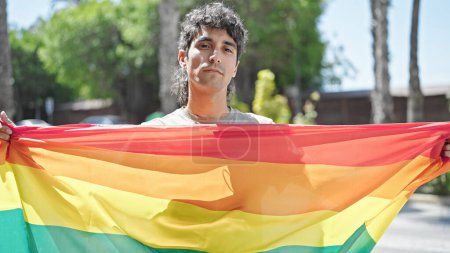 Foto de Joven hispano sosteniendo bandera de arco iris con expresión relajada en la calle - Imagen libre de derechos