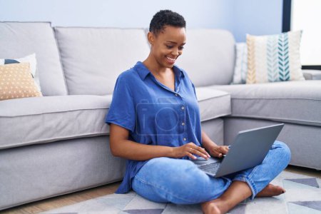 Téléchargez les photos : Femme afro-américaine utilisant un ordinateur portable assis sur le sol à la maison - en image libre de droit