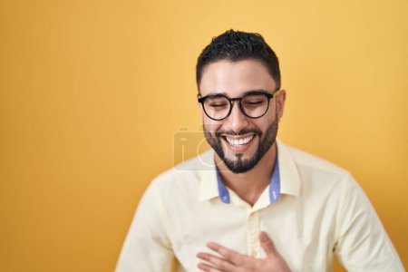 Téléchargez les photos : Jeune homme hispanique portant des vêtements de travail et des lunettes souriant et riant fort à haute voix parce que drôle blague folle avec les mains sur le corps. - en image libre de droit