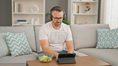 Photo for Grey-haired man eating salad using touchpad and headphones at home - Royalty Free Image