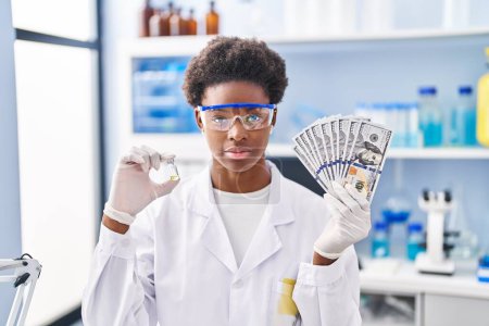 Foto de Mujer afroamericana trabajando en laboratorio científico sosteniendo dólares relajados con expresión seria en la cara. simple y natural mirando a la cámara. - Imagen libre de derechos