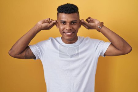 Téléchargez les photos : Jeune homme hispanique debout sur fond jaune souriant tirant les oreilles avec les doigts, geste drôle. problème d'audition - en image libre de droit