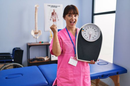 Photo for Young brunette woman as nutritionist holding weighing machine screaming proud, celebrating victory and success very excited with raised arms - Royalty Free Image