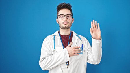 Téléchargez les photos : Jeune homme hispanique médecin faire un serment avec la main sur la poitrine sur fond bleu isolé - en image libre de droit