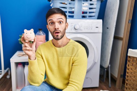 Photo for Hispanic man doing laundry holding piggy bank scared and amazed with open mouth for surprise, disbelief face - Royalty Free Image