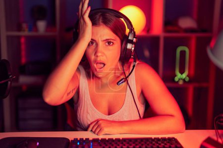 Photo for Young blonde woman playing video games wearing headphones surprised with hand on head for mistake, remember error. forgot, bad memory concept. - Royalty Free Image