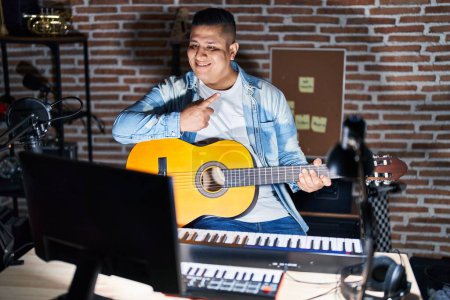 Téléchargez les photos : Jeune homme hispanique jouant de la guitare classique au studio de musique pointant du doigt vers le côté montrant la publicité, visage sérieux et calme - en image libre de droit