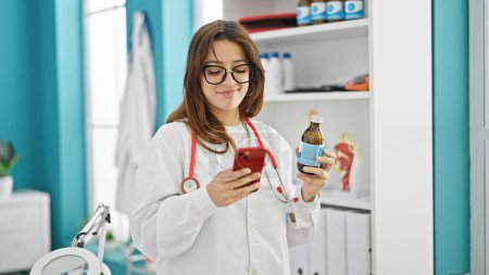 Photo for Young beautiful hispanic woman doctor using smartphone holding medication bottle at clinic - Royalty Free Image