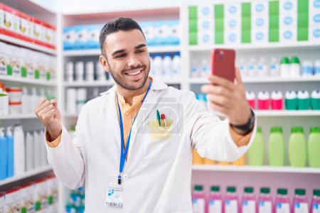 Téléchargez les photos : Jeune homme hispanique travaillant à la pharmacie drugstore faire appel vidéo avec smartphone crier fier, célébrer la victoire et le succès très excité avec le bras levé - en image libre de droit