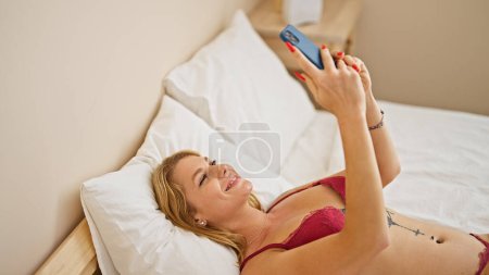 Photo for Young blonde woman wearing sensual lingerie lying on bed using smartphone at bedroom - Royalty Free Image