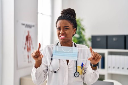 Photo for Young african american with braids wearing doctor uniform holding safety mask clueless and confused expression. doubt concept. - Royalty Free Image