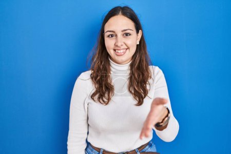 Foto de Mujer hispana joven de pie sobre fondo azul sonriendo amistoso ofreciendo apretón de manos como saludo y bienvenida. negocios exitosos. - Imagen libre de derechos