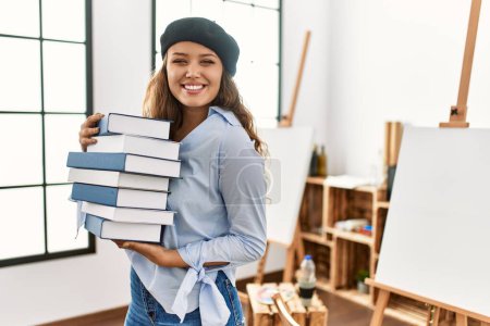 Photo for Young beautiful hispanic woman artist smiling confident holding books at art studio - Royalty Free Image