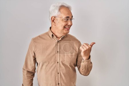 Foto de Hispanic senior man wearing glasses smiling with happy face looking and pointing to the side with thumb up. - Imagen libre de derechos