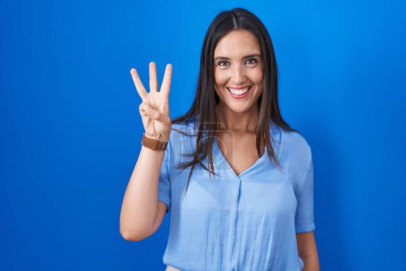 Photo for Young brunette woman standing over blue background showing and pointing up with fingers number three while smiling confident and happy. - Royalty Free Image