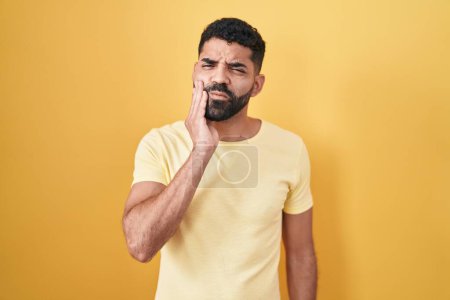 Foto de Hombre hispano con barba de pie sobre fondo amarillo tocando boca con mano con expresión dolorosa debido a dolor de muelas o enfermedad dental en los dientes. dentista - Imagen libre de derechos