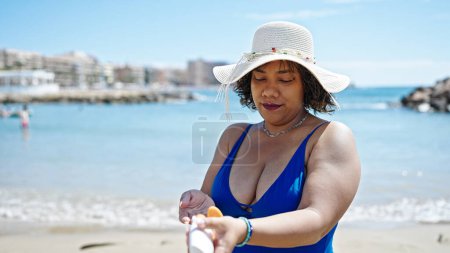 Foto de Joven hermosa turista latina usando traje de baño y sombrero de verano aplicando protector solar en el brazo en la playa - Imagen libre de derechos