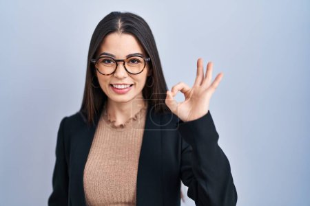 Foto de Young brunette woman standing over blue background smiling positive doing ok sign with hand and fingers. successful expression. - Imagen libre de derechos