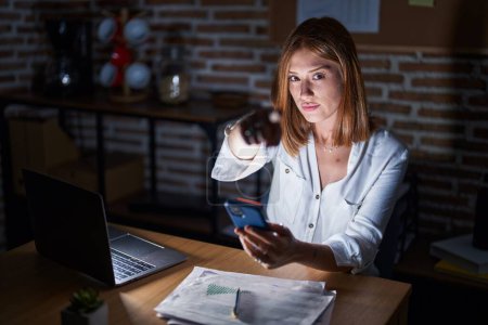 Téléchargez les photos : Jeune femme rousse travaillant au bureau la nuit pointant du doigt la caméra et vers vous, geste confiant et sérieux - en image libre de droit