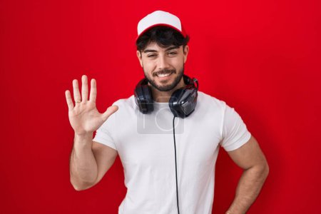 Téléchargez les photos : Homme hispanique avec barbe portant chapeau de joueur et écouteurs montrant et pointant vers le haut avec les doigts numéro cinq tout en souriant confiant et heureux. - en image libre de droit