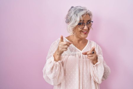 Foto de Middle age woman with grey hair standing over pink background pointing fingers to camera with happy and funny face. good energy and vibes. - Imagen libre de derechos