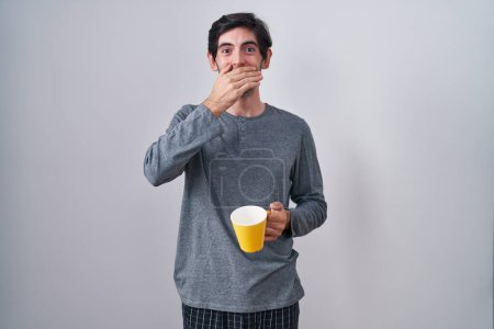 Foto de Un joven hispano con pijama bebiendo una taza de café sorprendió cubriendo la boca con las manos por error. concepto secreto. - Imagen libre de derechos