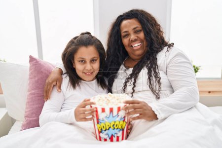 Photo for Mother and young daughter eating popcorn in the bed smiling with a happy and cool smile on face. showing teeth. - Royalty Free Image